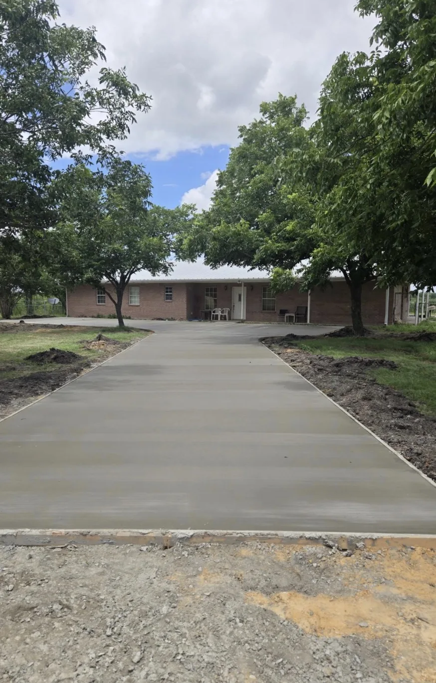 Freshly poured concrete driveway leading to a single-story brick house under shade trees